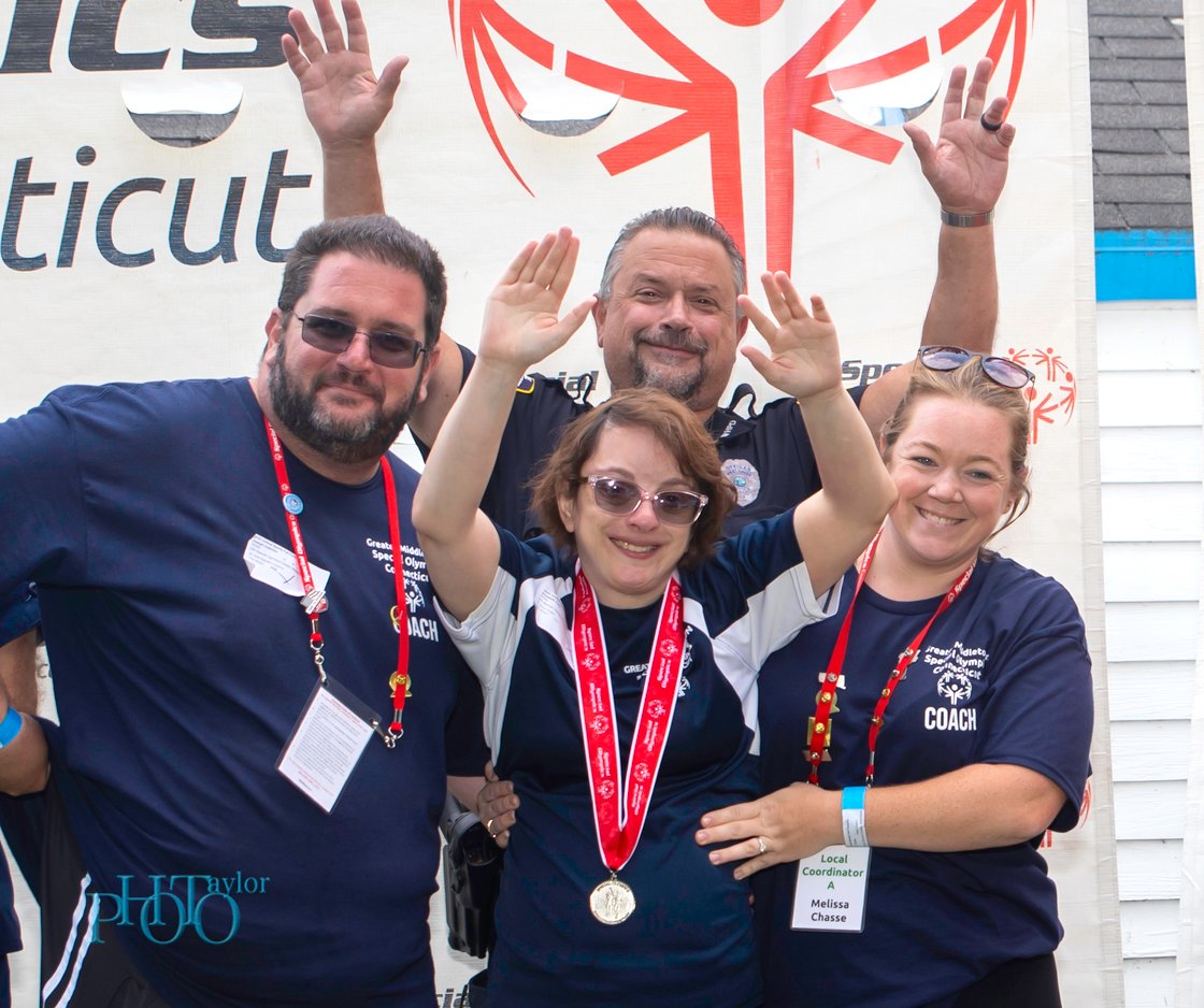 Four people pose for picture, athlete with medal in middle with arms up